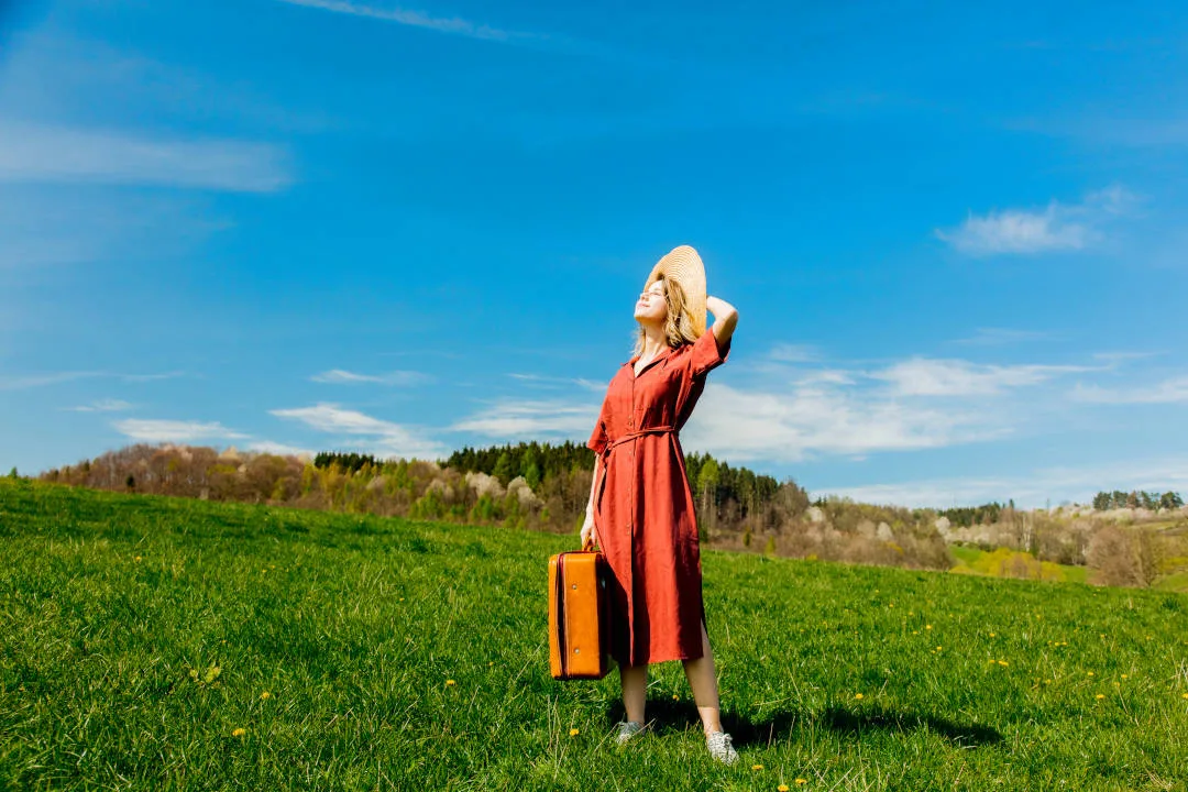 Beautiful girl in red dress and hat with suitcase on meadow. Spring season